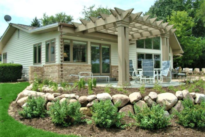 House with stone patio, pergola, and white outdoor chairs in a landscaped garden