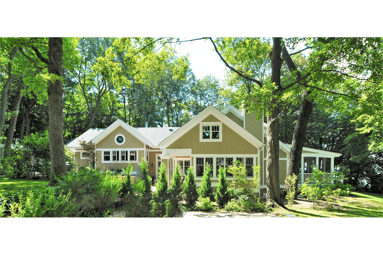 Small beige house with gabled roofs surrounded by tall green trees and lush landscaping
