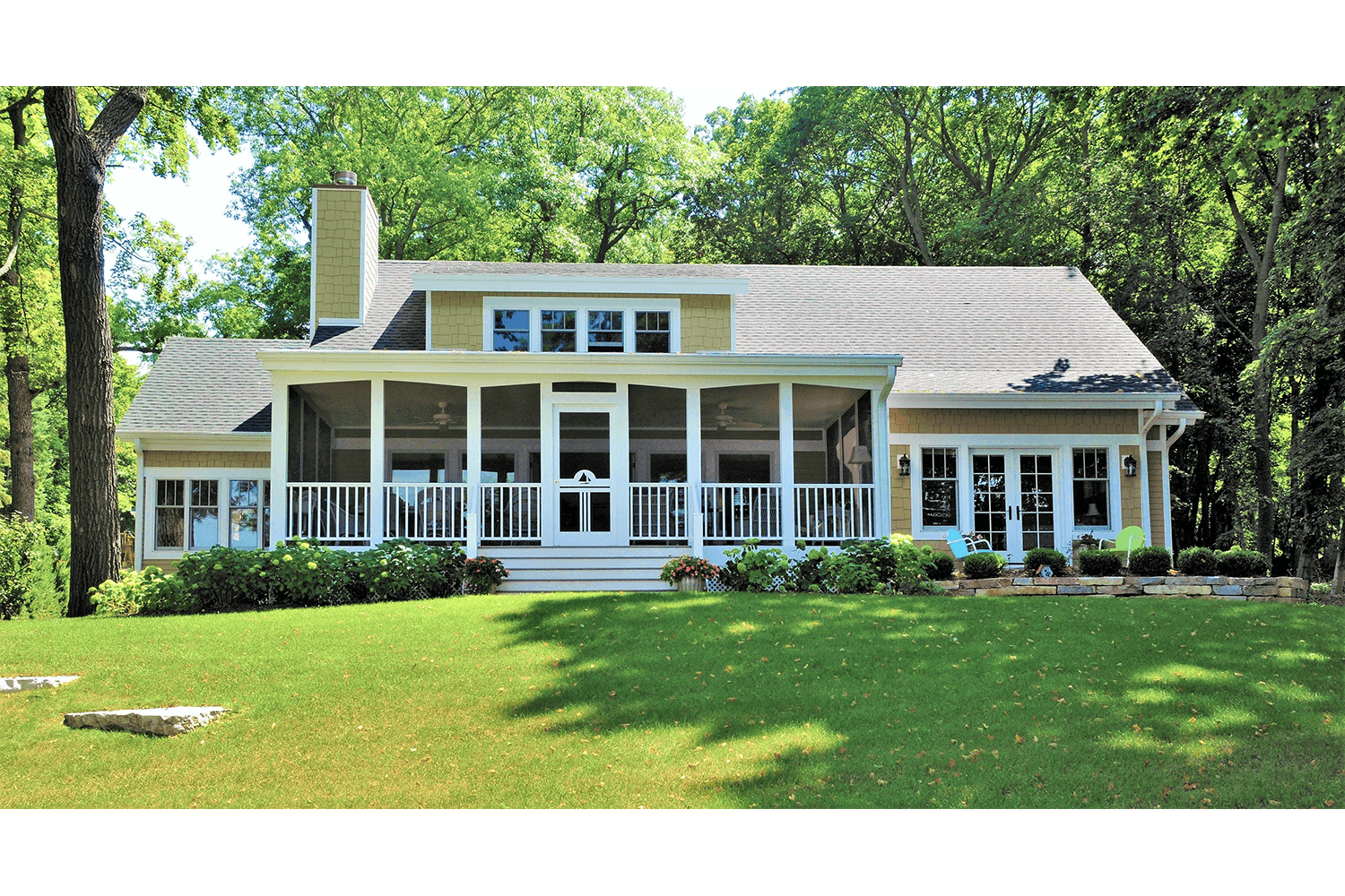 White house with a large front porch and green lawn, surrounded by trees in daylight