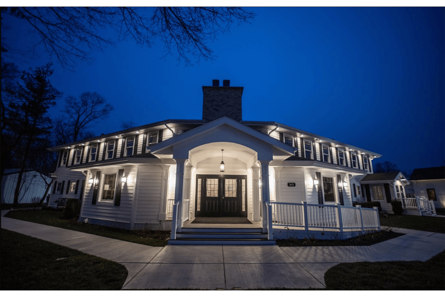 Night view of a lit white house with a central porch and chimney, seen from the front walkway