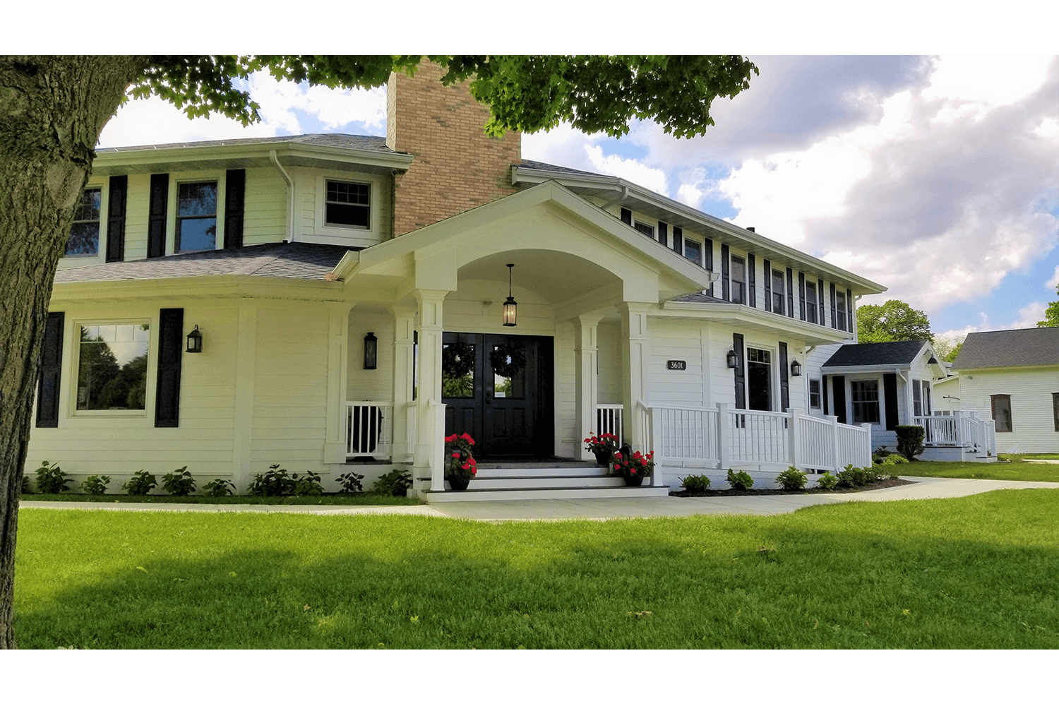 Large white Victorian house with wraparound porch and green lawn under a partly cloudy sky