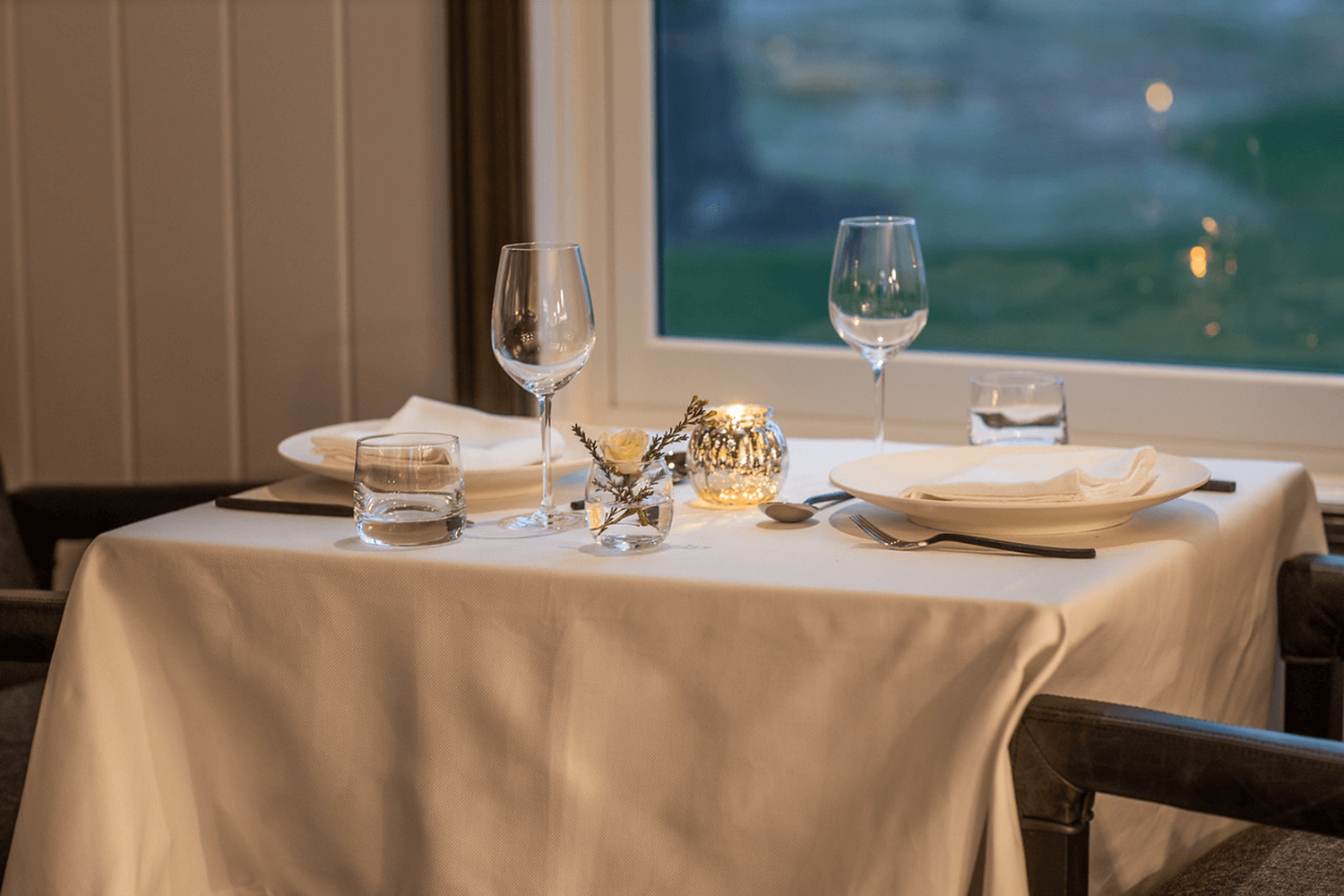Set dining table by a window with white tablecloth, wine glasses, plates, and candlelight