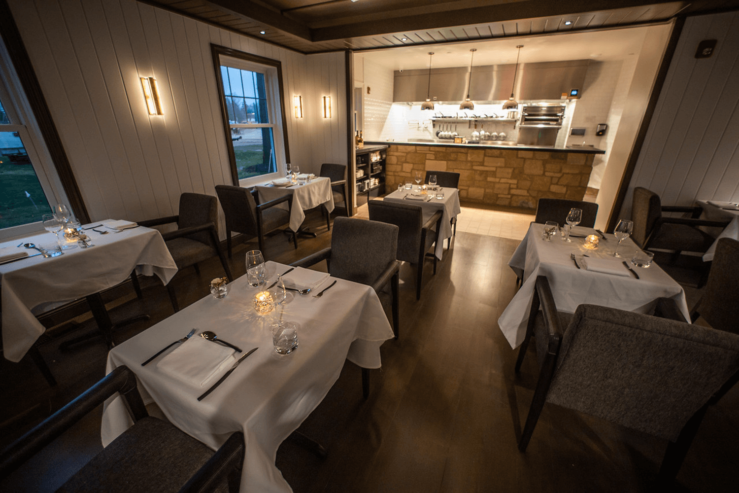 Empty restaurant dining room with white tablecloths and a lit open kitchen in back