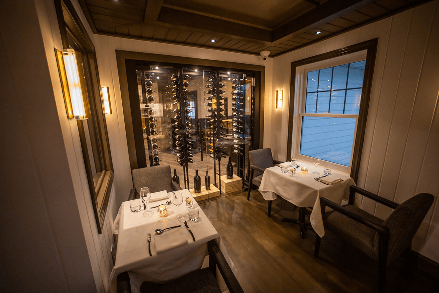Dimly lit elegant dining room with white tablecloths, set tables, and a window view outside.