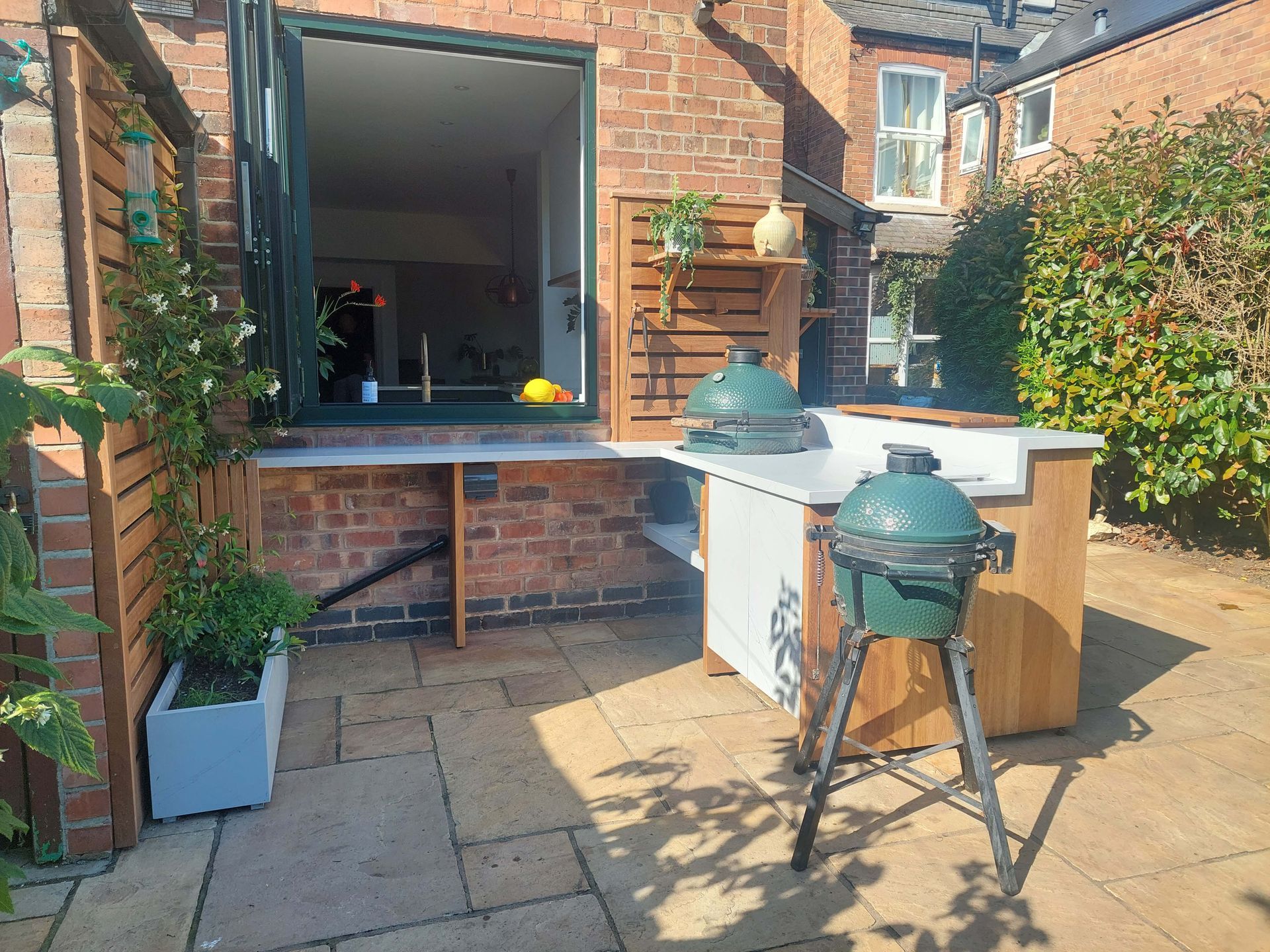 Backyard patio with brick wall, open window, potted plants, and a green barbecue grill on a tripod