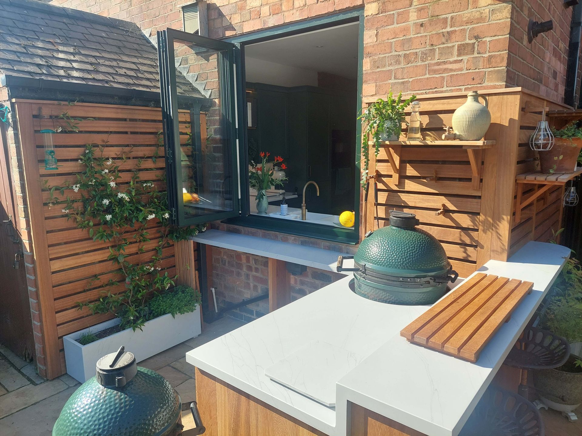 Backyard patio with open window, brick wall, wooden planter, and a white table with green dome-shaped grill.