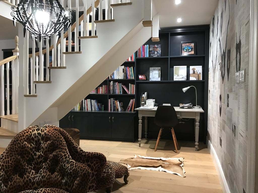 Leopard lying on a living room floor beneath stairs near a desk and shelving unit