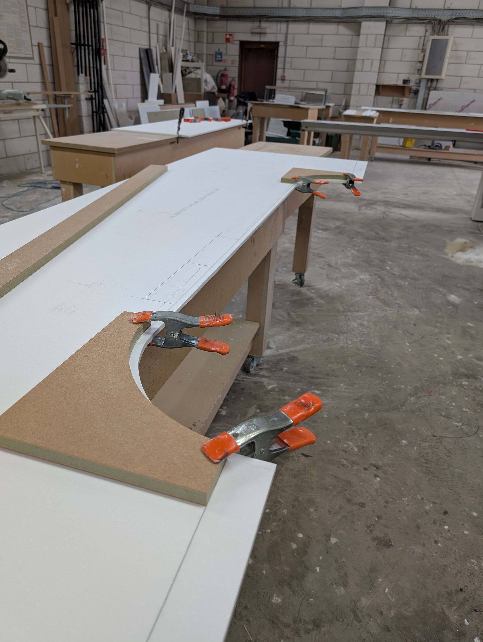 Curved white board clamped to a workbench in a workshop, with orange clamps on the edge
