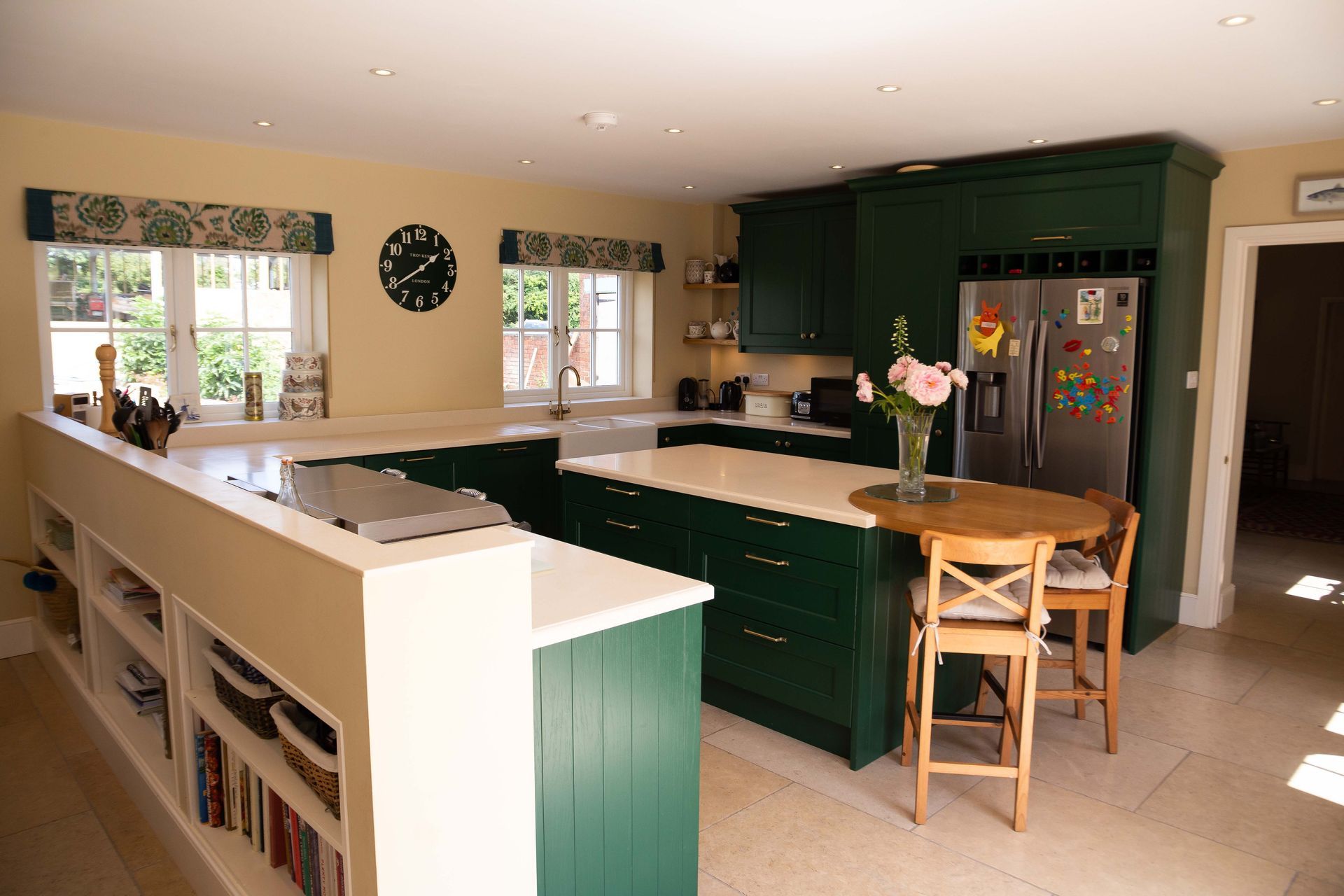 Bright kitchen with dark green cabinets, white counters, barstools, and a stainless steel refrigerator.