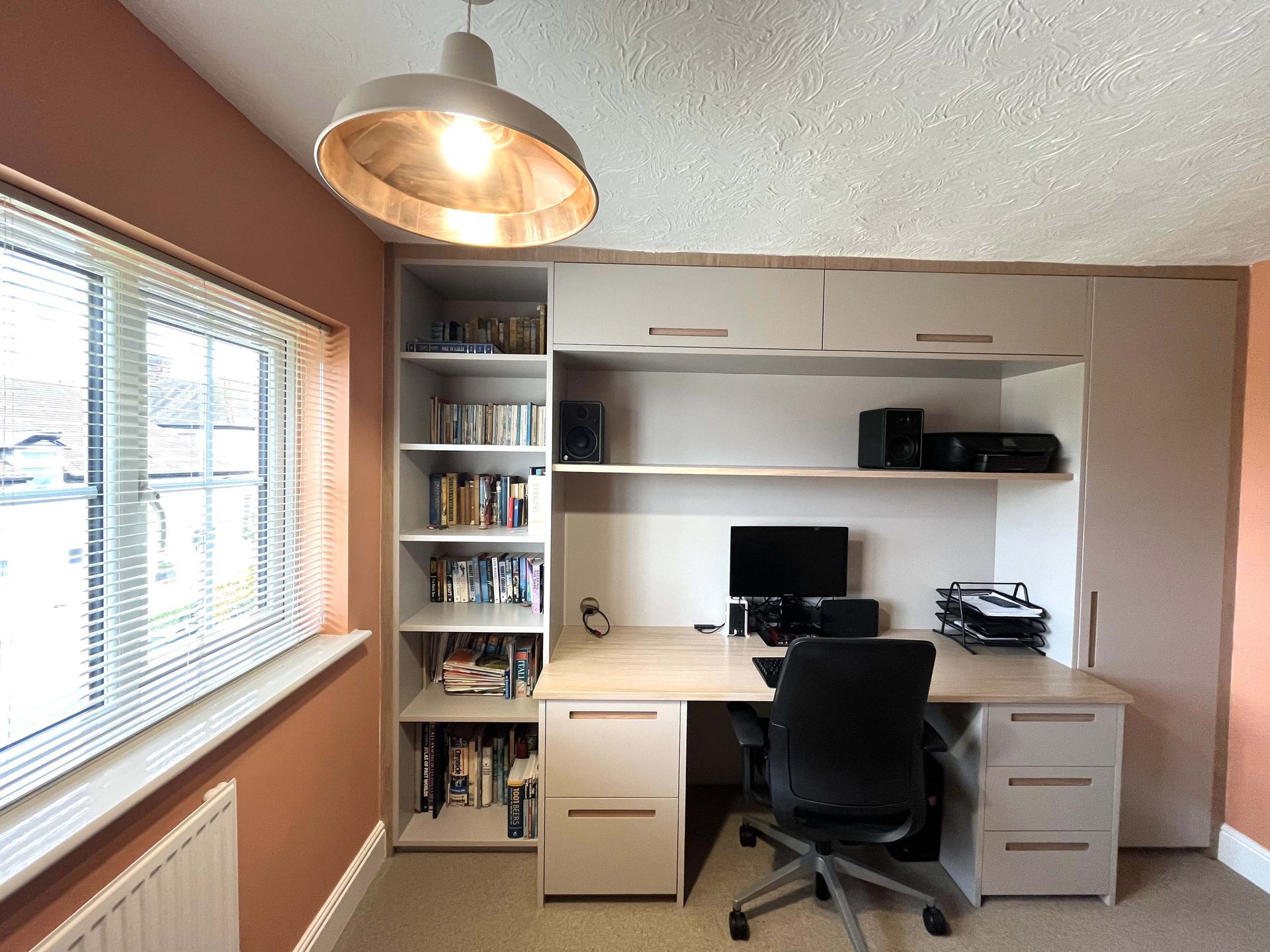Home office with desk, black chair, shelves, computer, and large window in a beige room