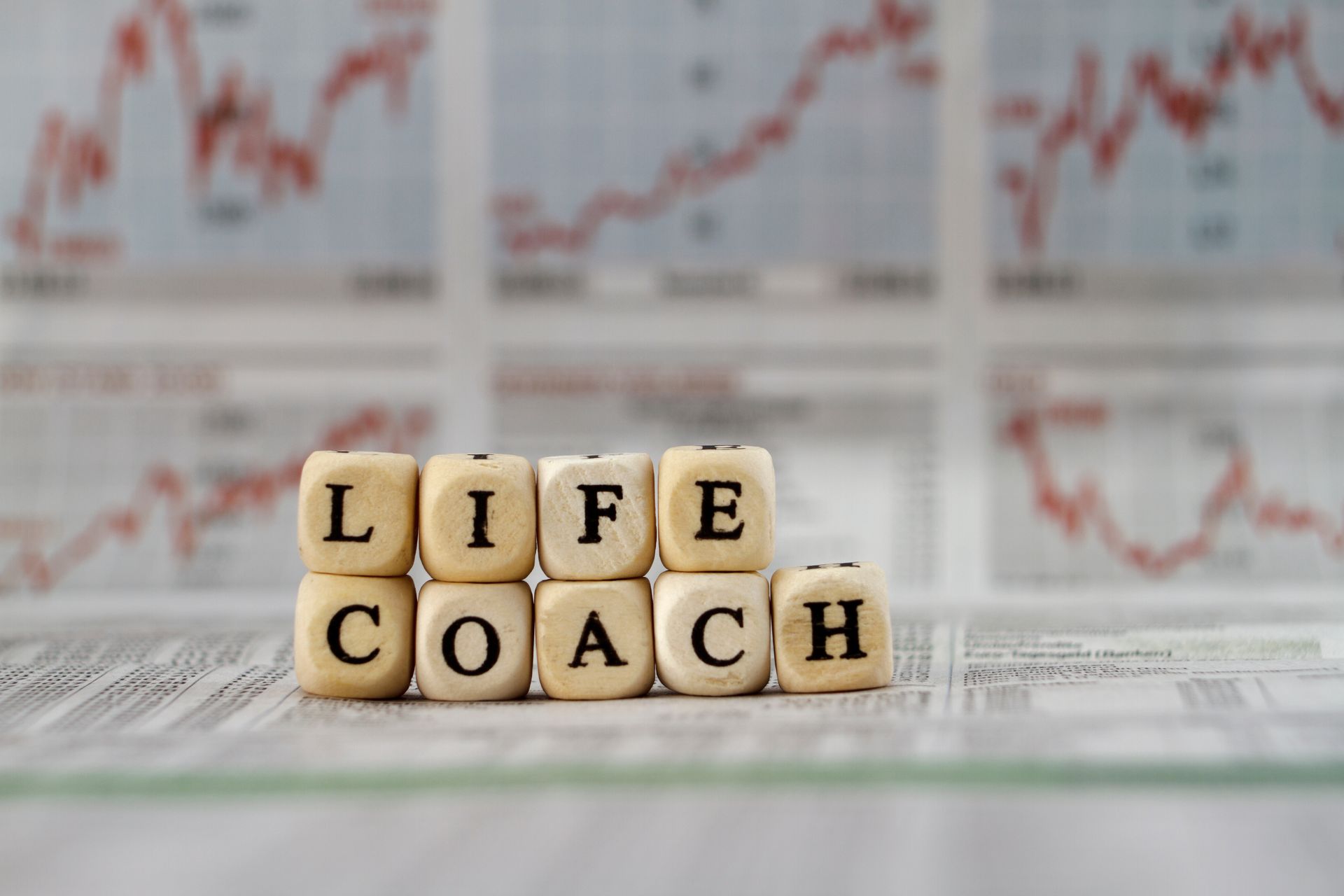 Wooden blocks spelling “LIFE COACH” on newspaper, with blurry stock charts in the background