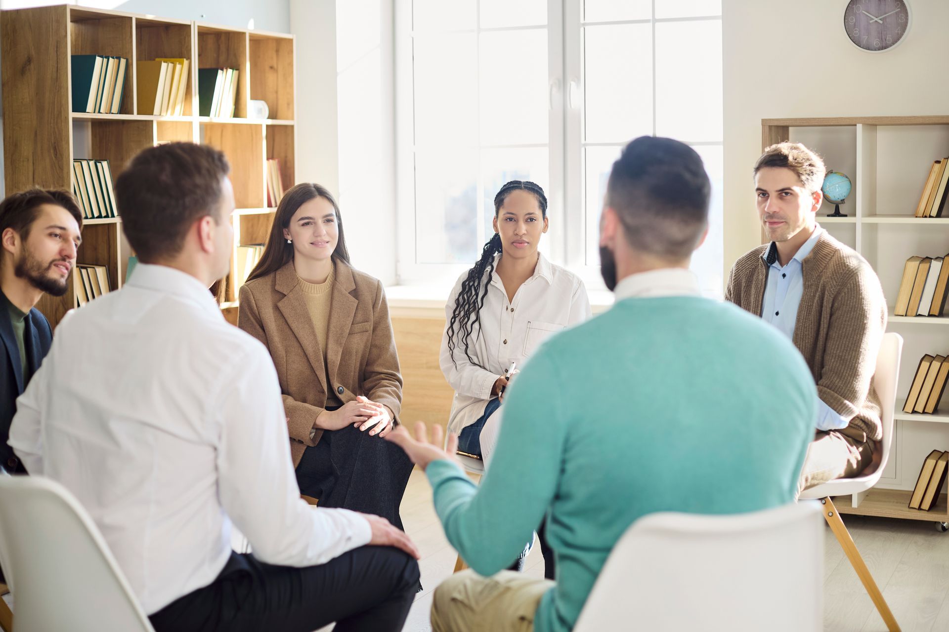 Group seated in a bright office discussing in a circle, with shelves and a clock in the background.