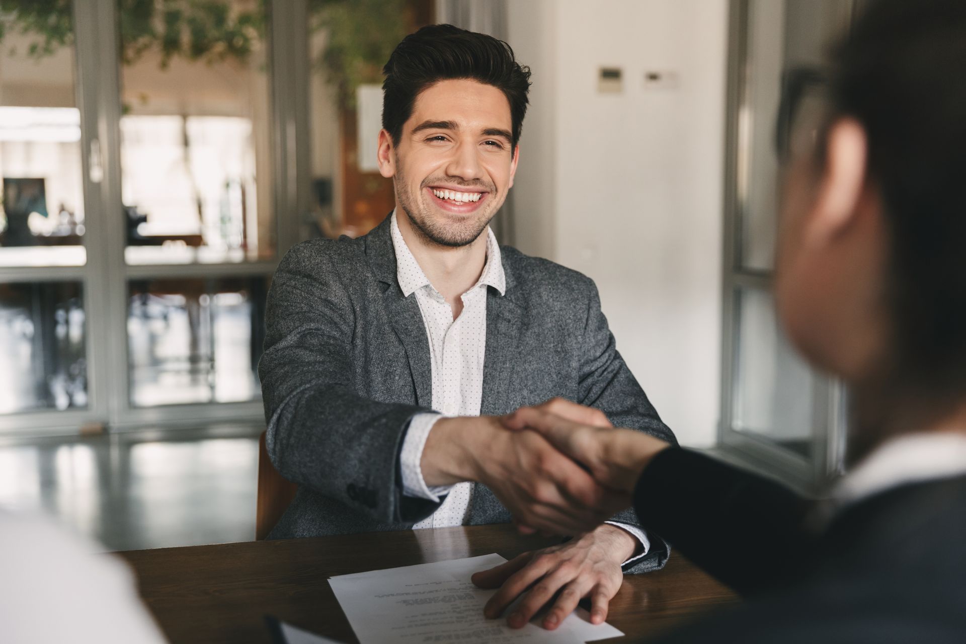 Two people shaking hands across a table in a bright office, with one smiling in the foreground