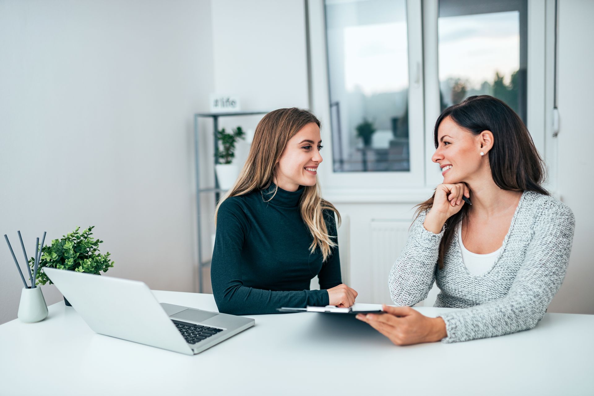 Two women talking at a desk with a laptop in a bright office