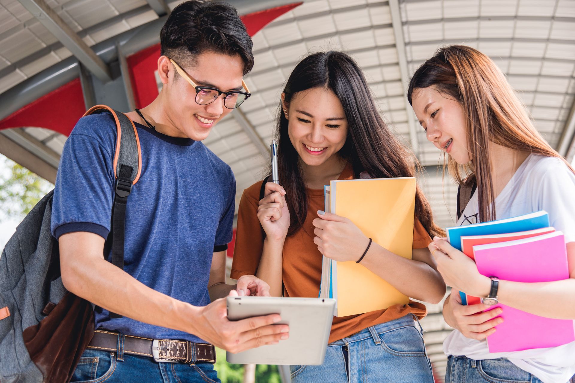 Three students looking at a tablet and smiling, carrying notebooks and backpacks in a hallway