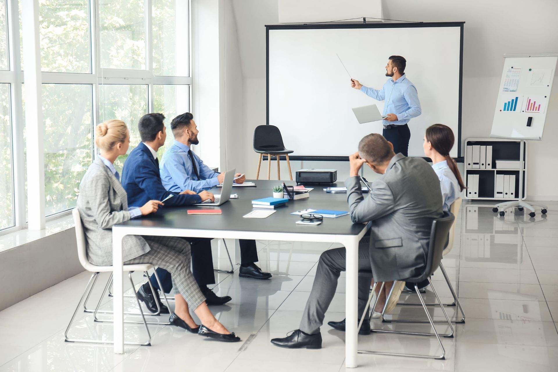 Business meeting in a bright conference room, with a presenter pointing at a whiteboard and attendees seated at a table.