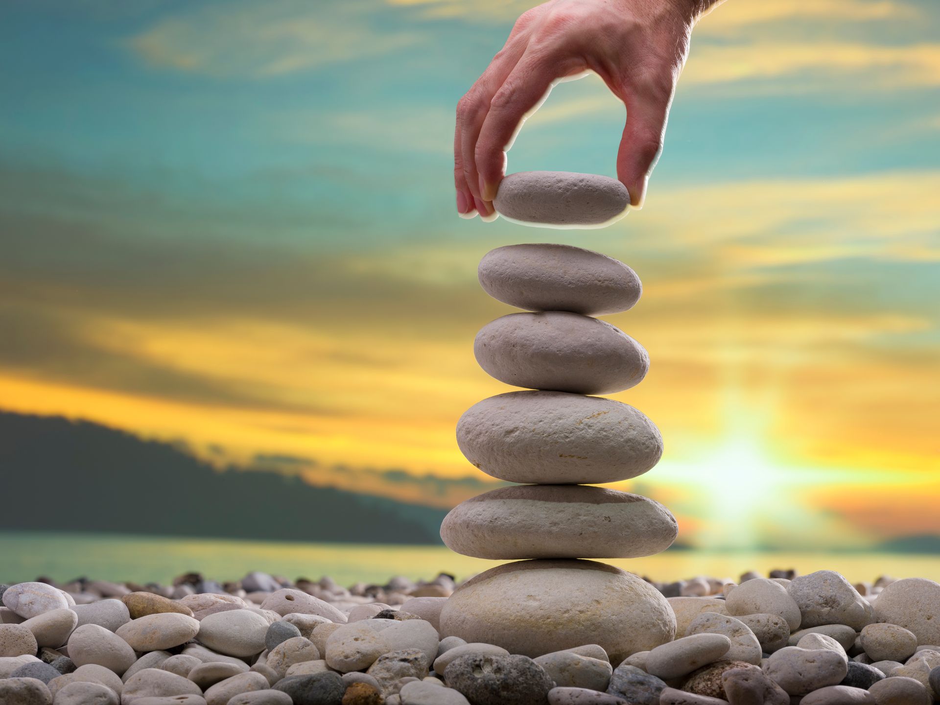 Hand stacking smooth stones on a balanced pebble tower at sunset by the water