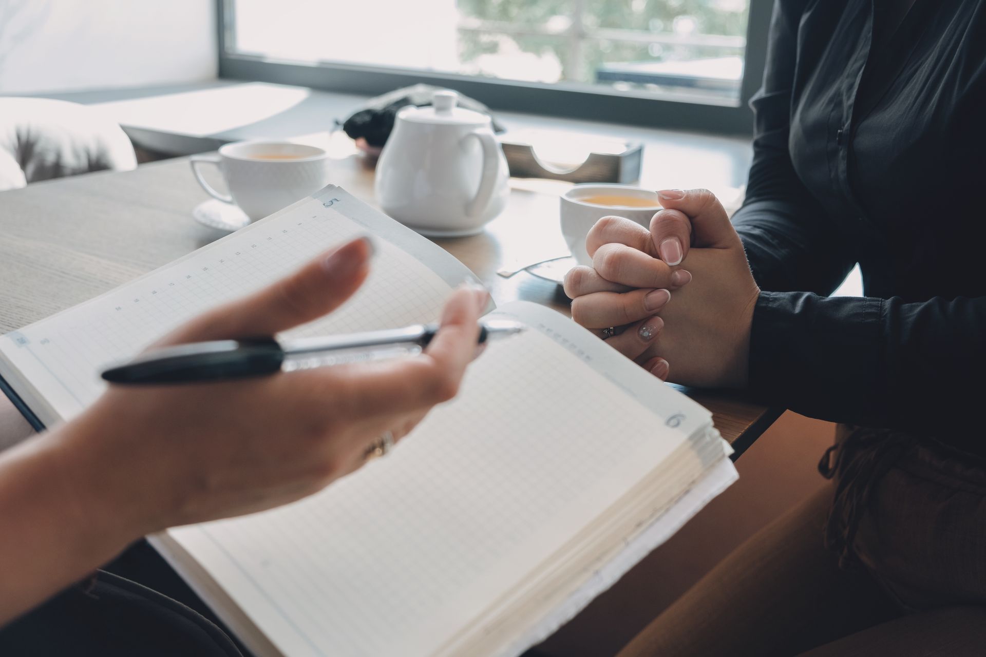 Two people talking at a desk, one taking notes in a notebook beside a teapot and coffee cup.