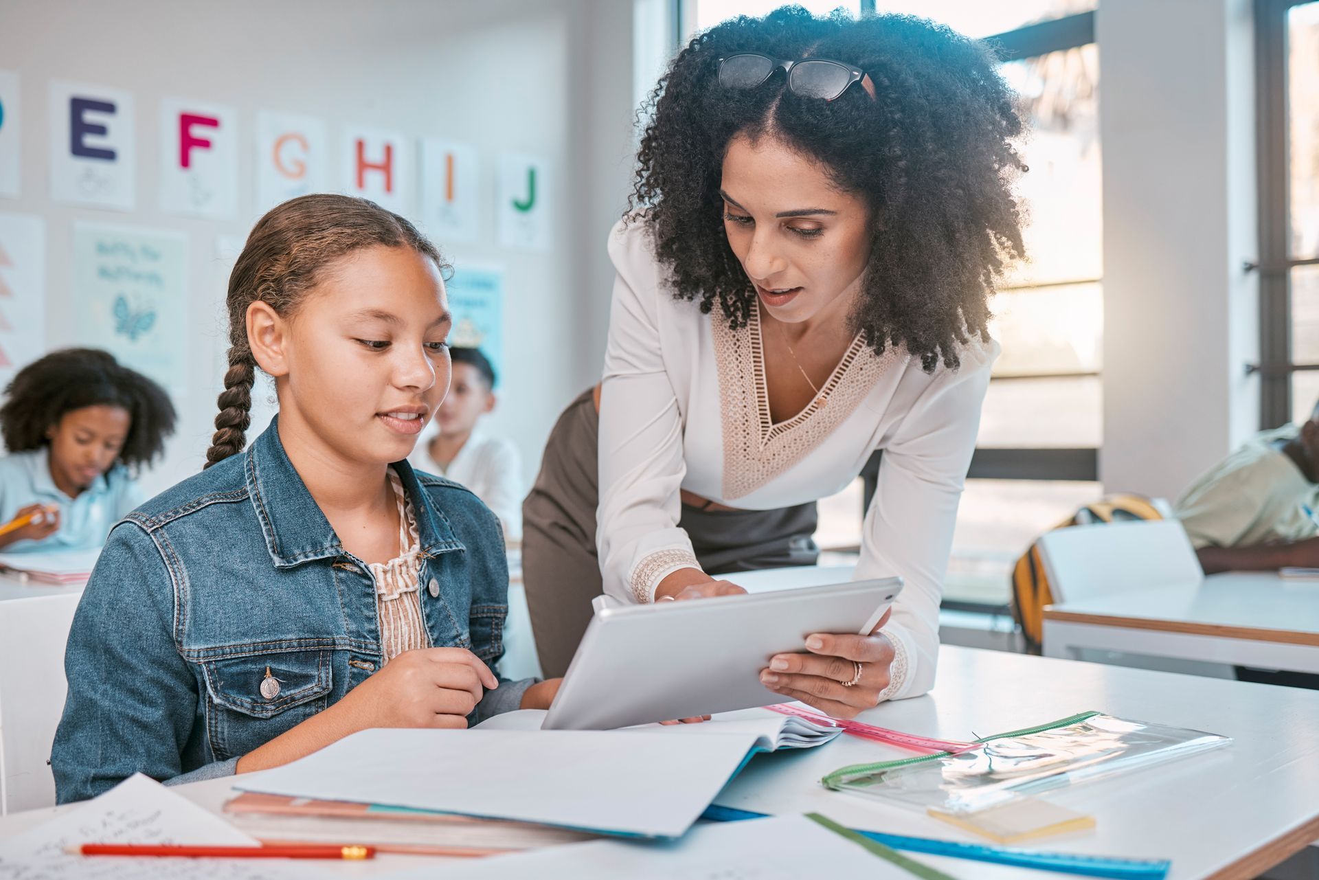 Teacher helping a student with a tablet at a classroom desk, with notebooks and alphabet letters on the wall
