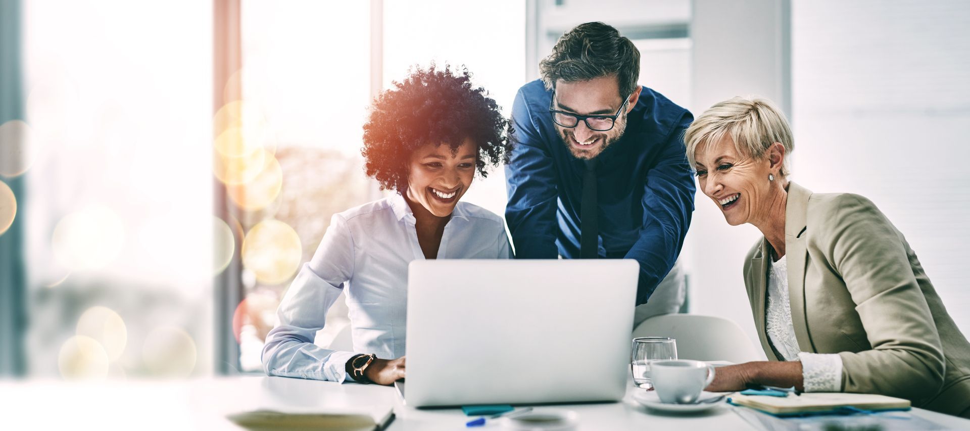 Three colleagues smiling around a laptop in a bright office meeting