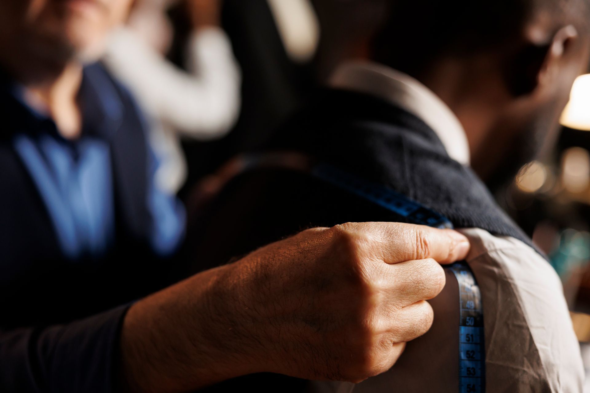 Hand measuring a tailor’s shoulder seam with a blue tape measure, with others blurred in the background