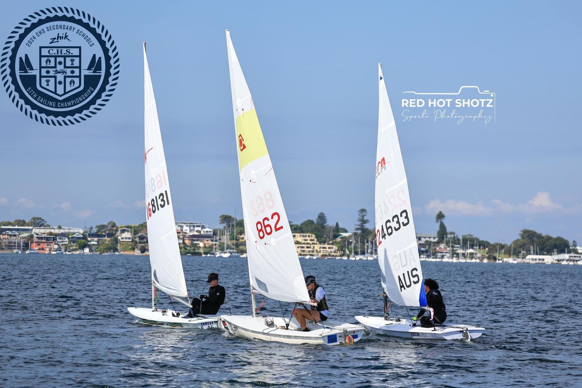 Three sailboats racing on a lake. White sails, black hulls, people aboard. Buildings and trees in the background. - Gosford Sailing Club in Gosford NSW