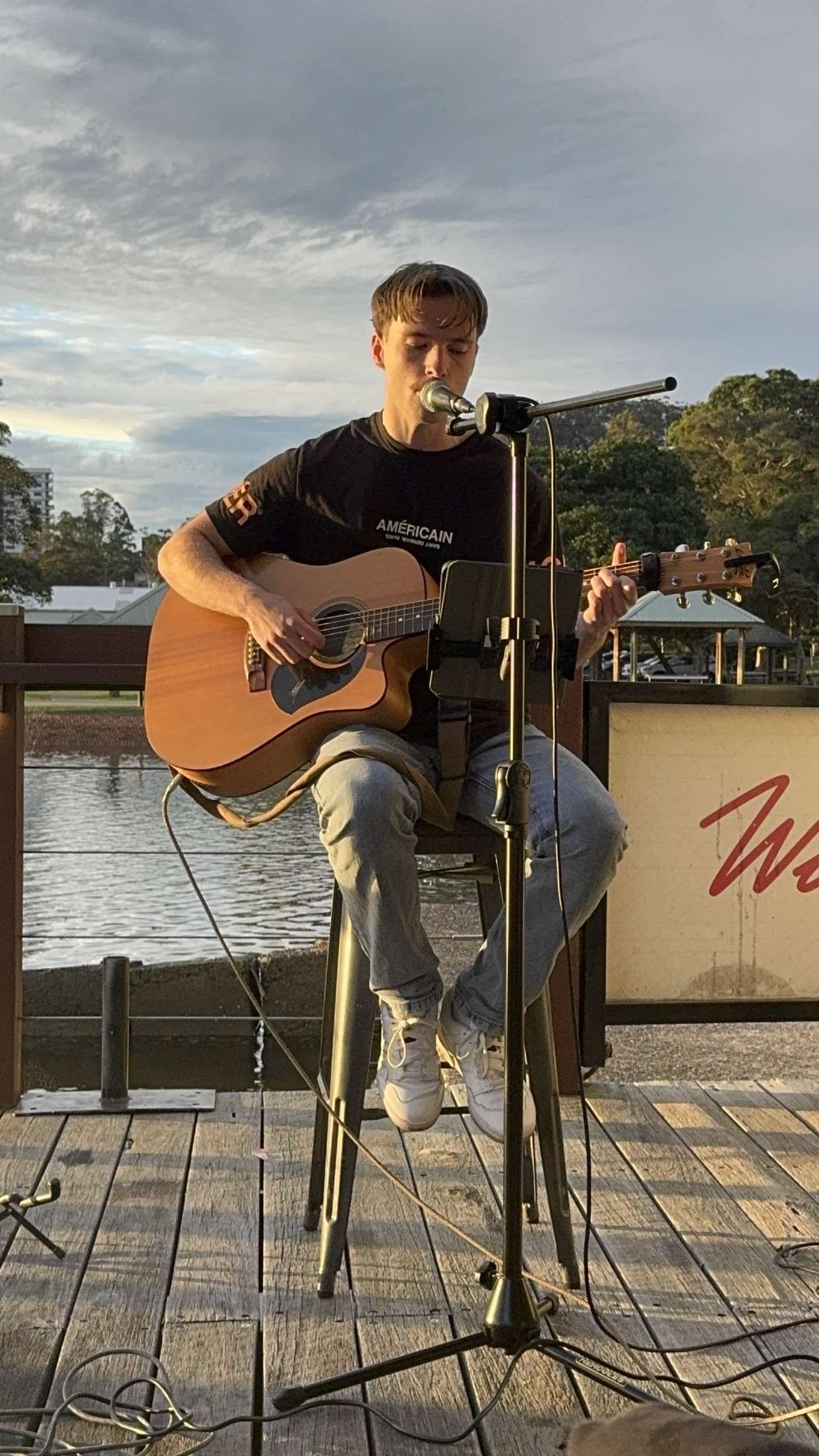 Person playing acoustic guitar on a stool by the waterfront at sunset — Gosford Sailing Club in Gosford, NSW