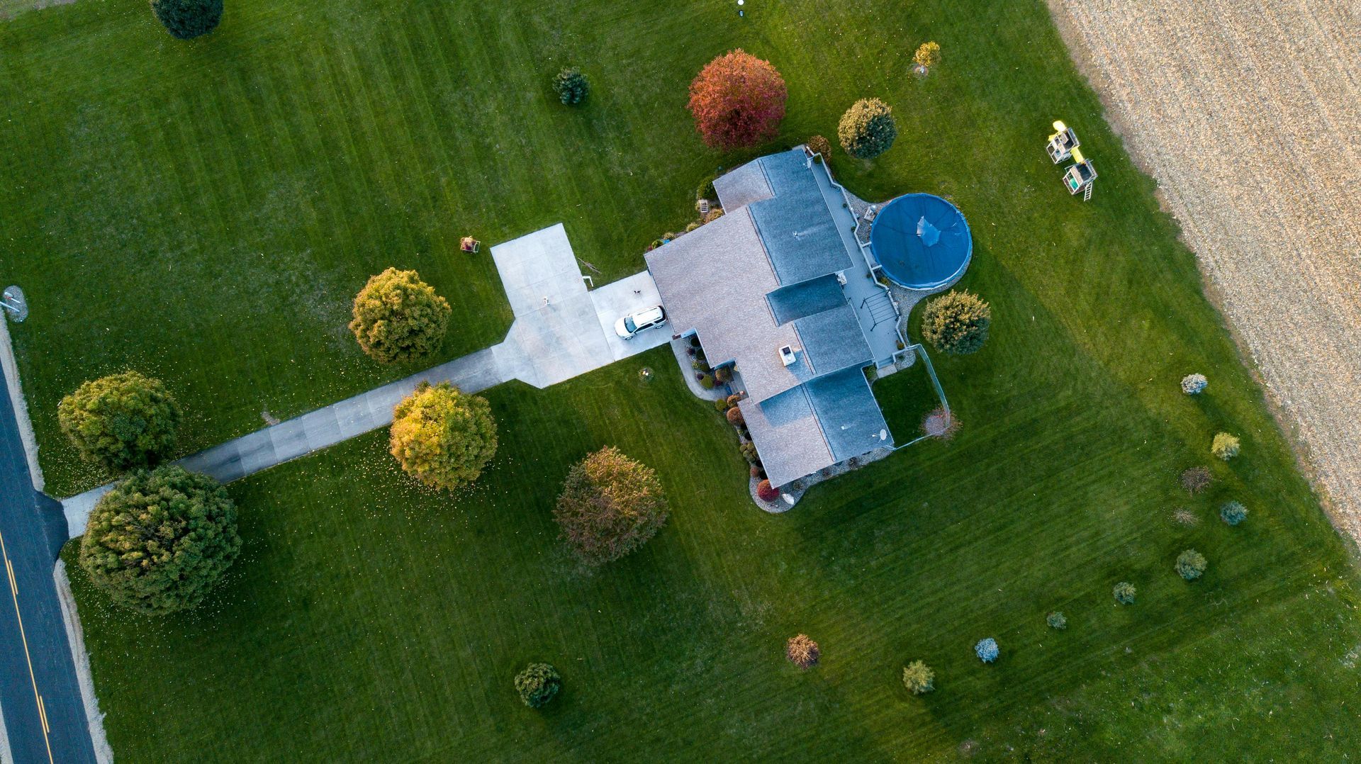 Aerial view of a house with a blue pool and green lawn, with trees and a driveway.