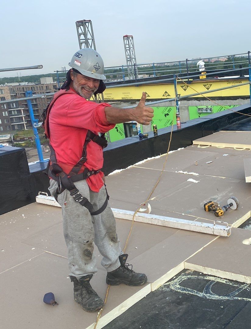 Ouvrier du bâtiment sur un toit, pouce levé. Il porte un harnais de sécurité, une chemise rouge, un pantalon gris et un casque de chantier.