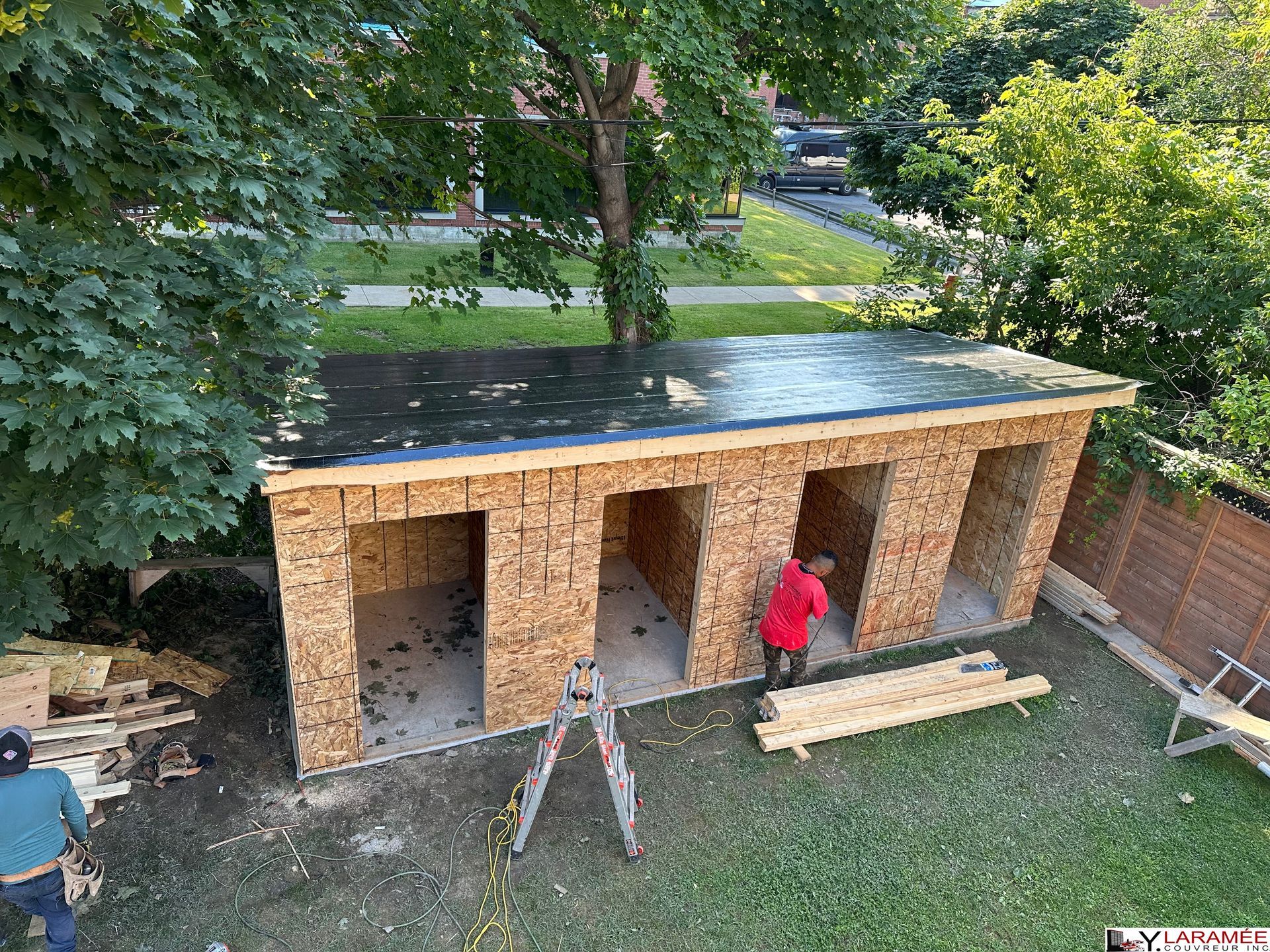 Construction of a multi-bay shed in a backyard; people working on the walls, roof, and interior.