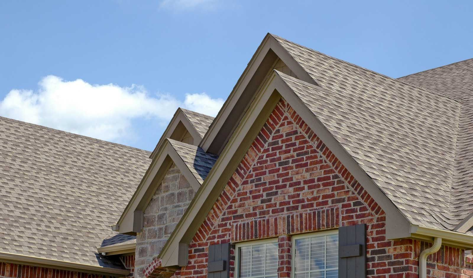Brick house with brown shingled roof, two gables, and blue sky.