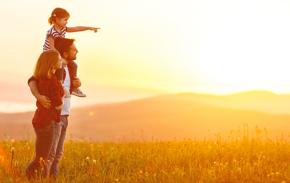 Family watching sunset; father carrying child on shoulders, mother beside them, golden field.