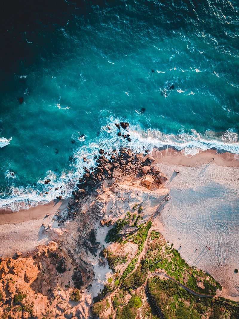 Aerial view of a beach with teal ocean waves crashing on brown rocks and sandy shore, green cliff on the right.