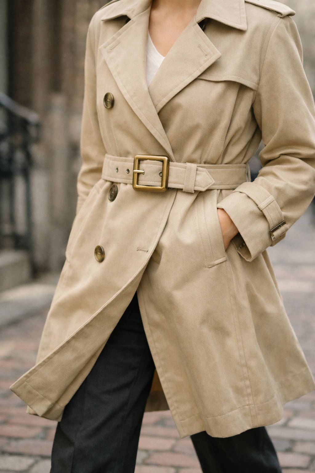 Person wearing a beige trench coat with belt, standing outdoors on cobblestones.