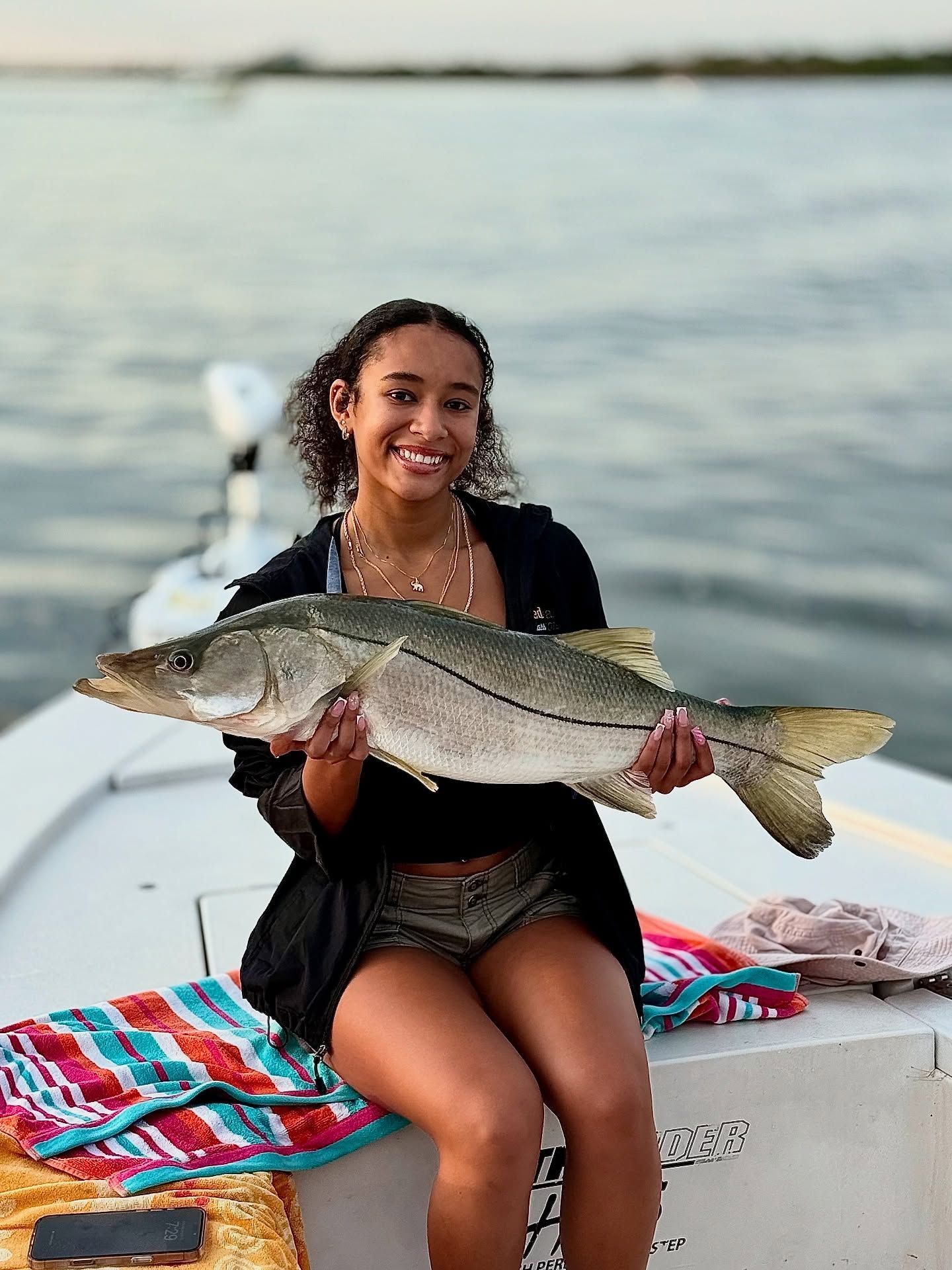 Woman smiling, holding a large fish on a boat. Water and sky in background.