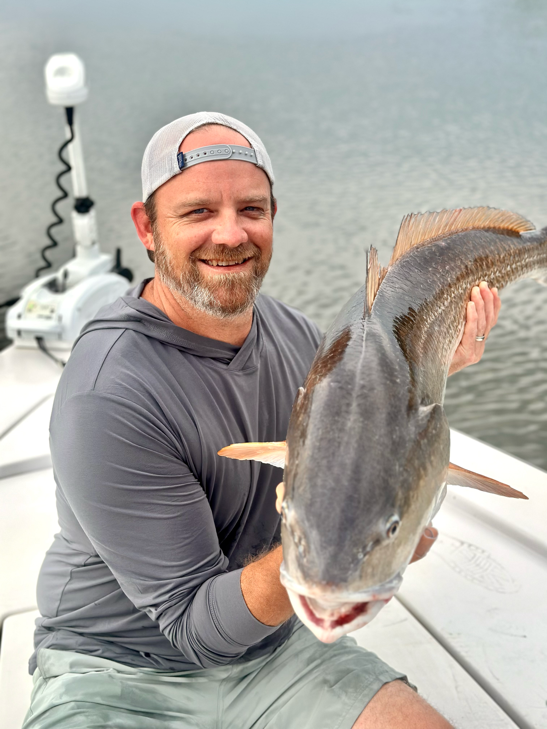 Man on a boat holds up a large red drum fish with a happy expression; overcast day.