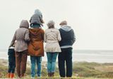 A group of people wearing jackets and coats standing in a line at the beach, looking out toward the water.