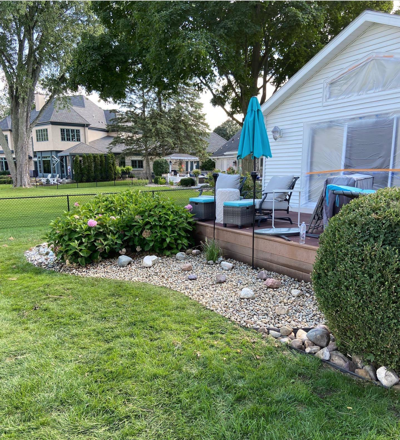 A white siding house with a wood deck, patio furniture, and a blue umbrella, overlooking a lawn with a rock garden.