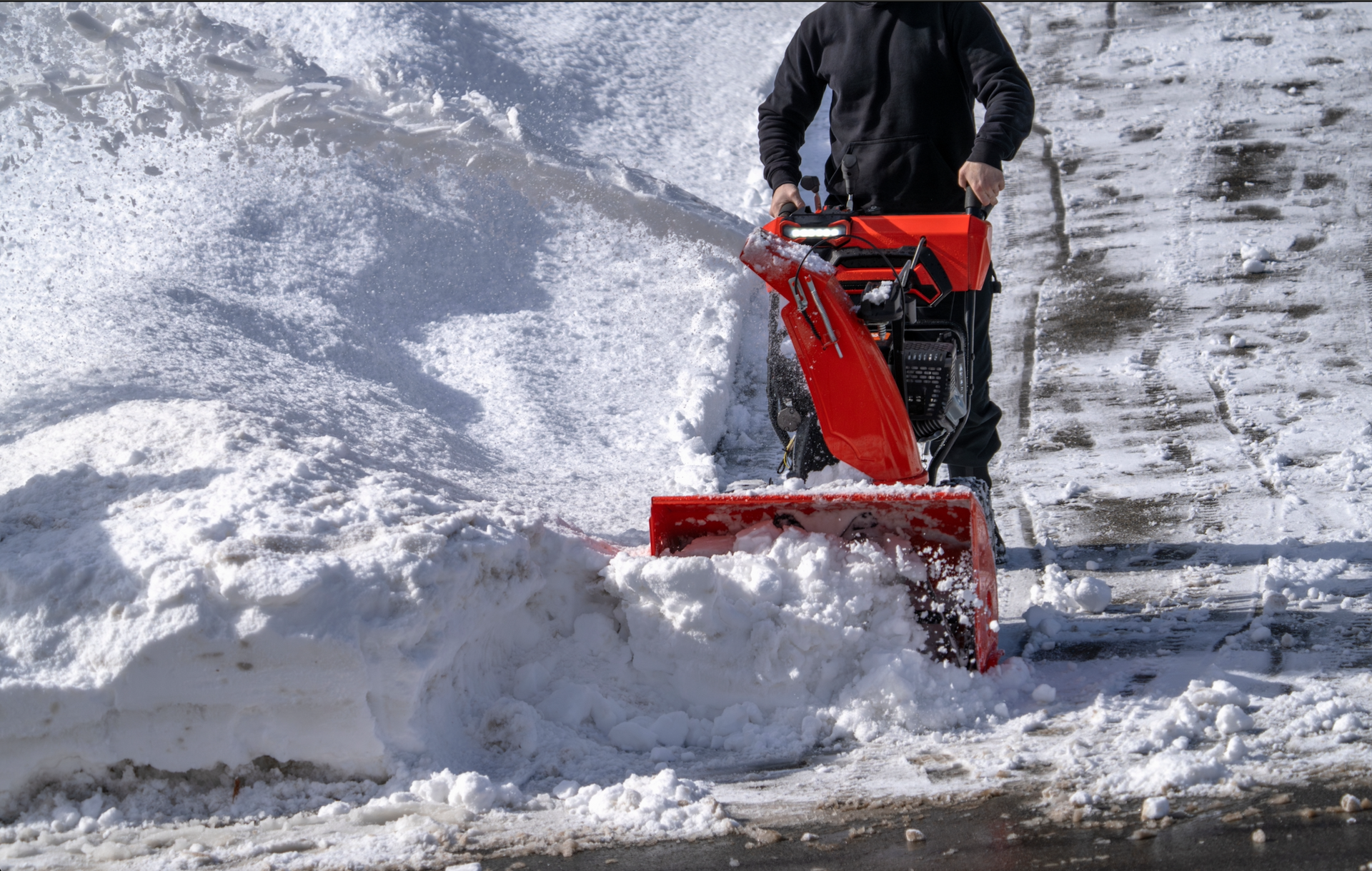 A person in a black jacket uses a bright orange snowblower to clear a deep pile of snow on a walkway.