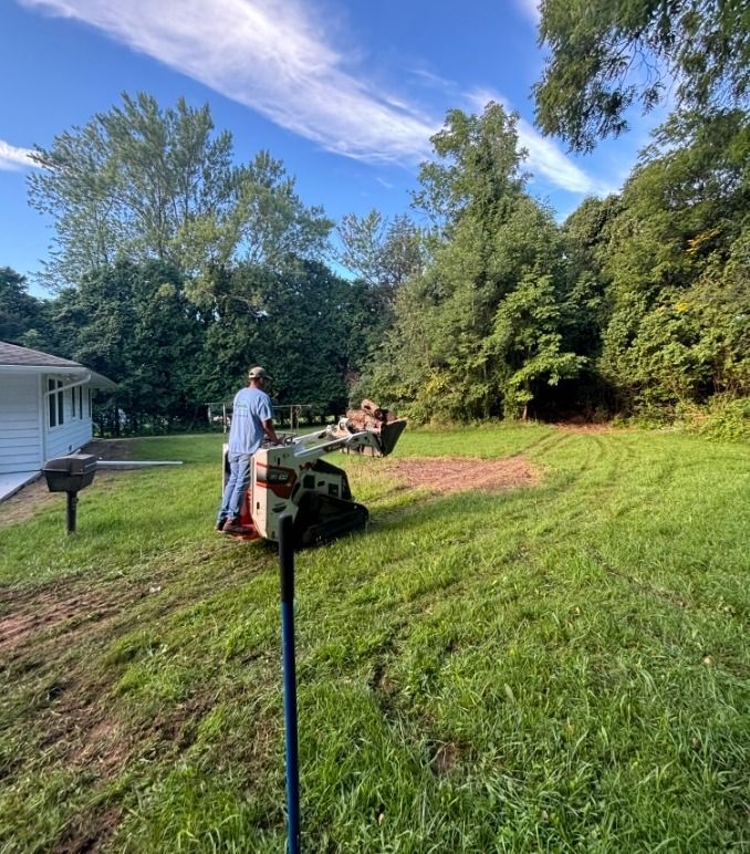 Person mowing a grassy yard beside a white house, with trees and a blue sky in the background.
