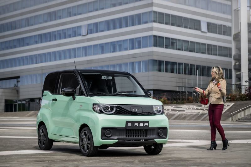 Un'auto compatta verde menta è parcheggiata accanto a un moderno edificio per uffici, con una persona in piedi accanto.