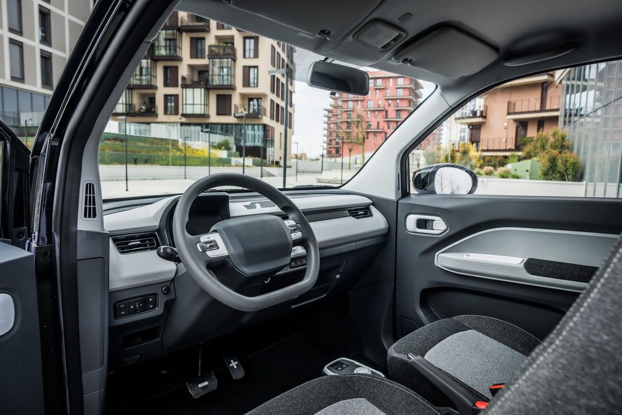 Interno di un'auto con volante e cruscotto, parcheggiata accanto a dei palazzi in una via cittadina.