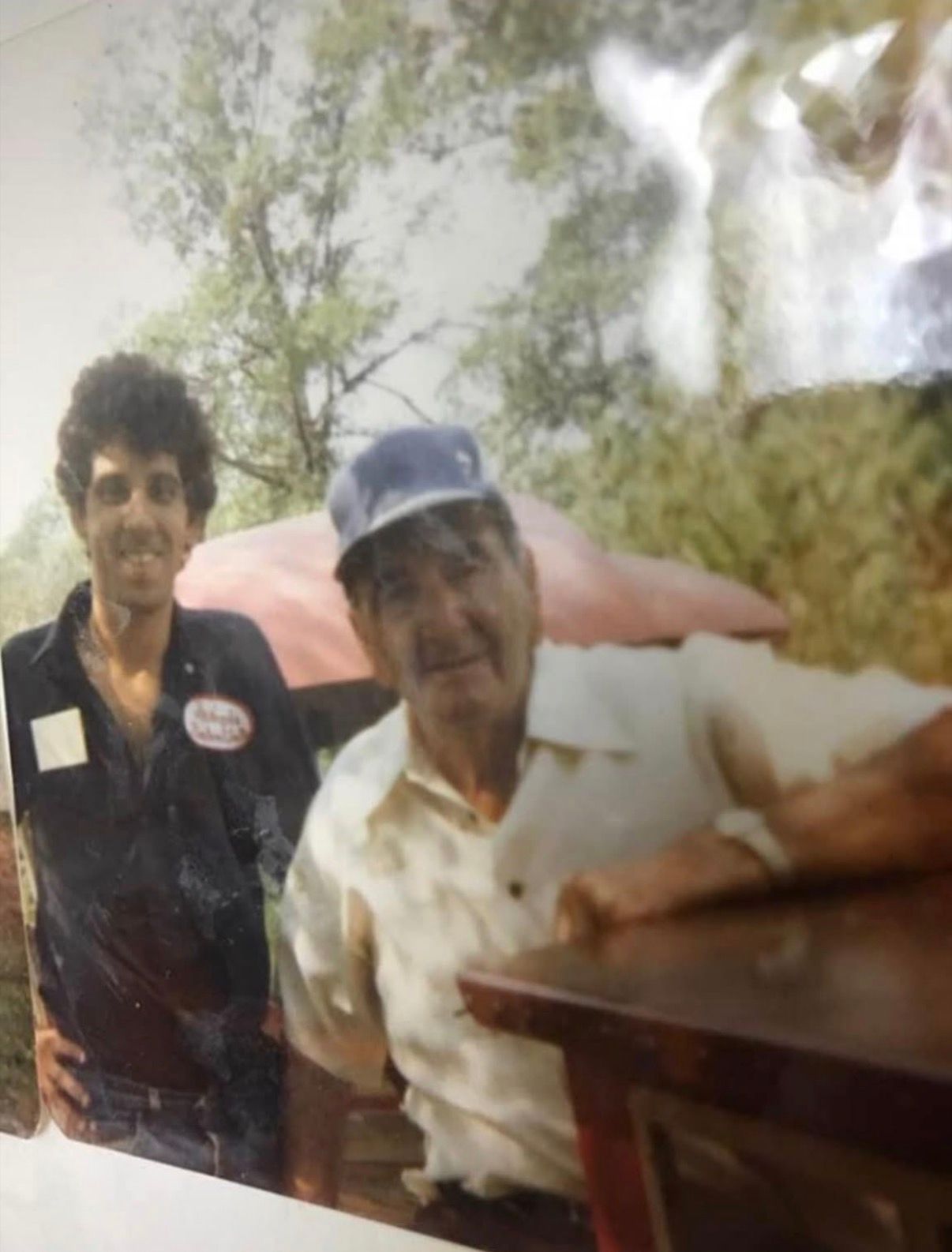 Two people outdoors by a table, one in a blue cap and white shirt, with trees in the background.