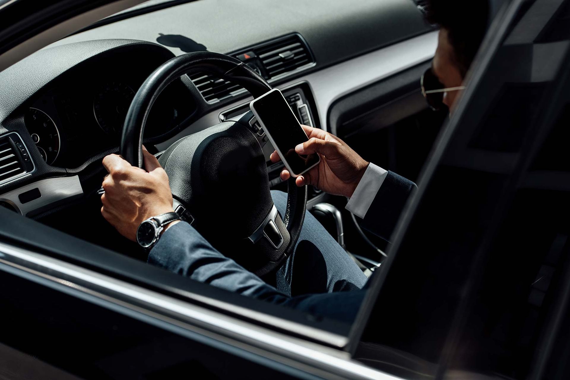 Persona in giacca e cravatta che usa il telefono mentre guida un'auto; luce solare visibile attraverso il finestrino.