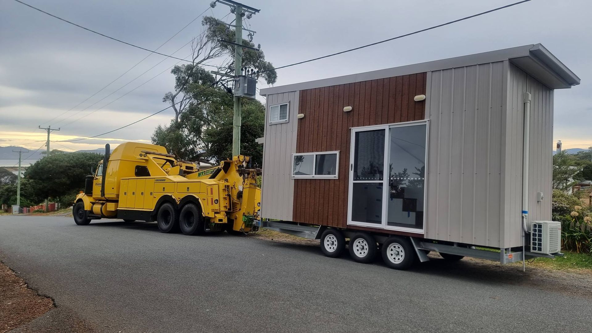 A yellow tow truck is towing a tiny house on a trailer.