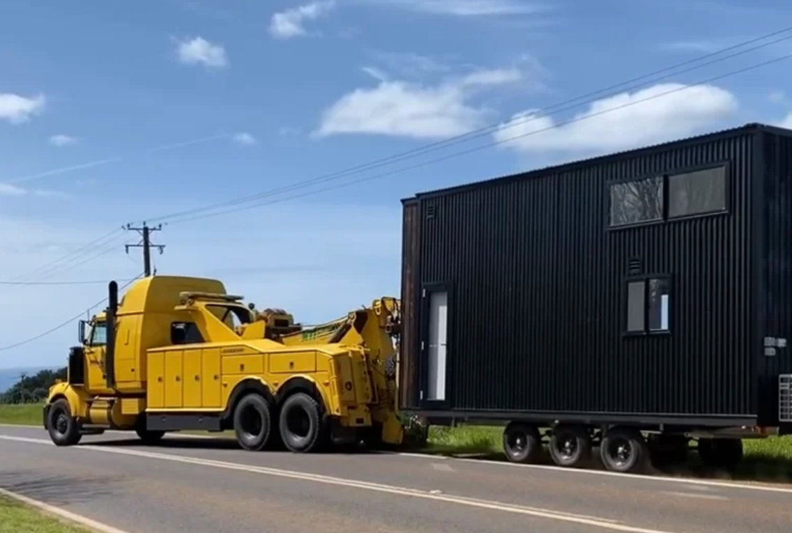 A yellow tow truck is towing a tiny house down the road.