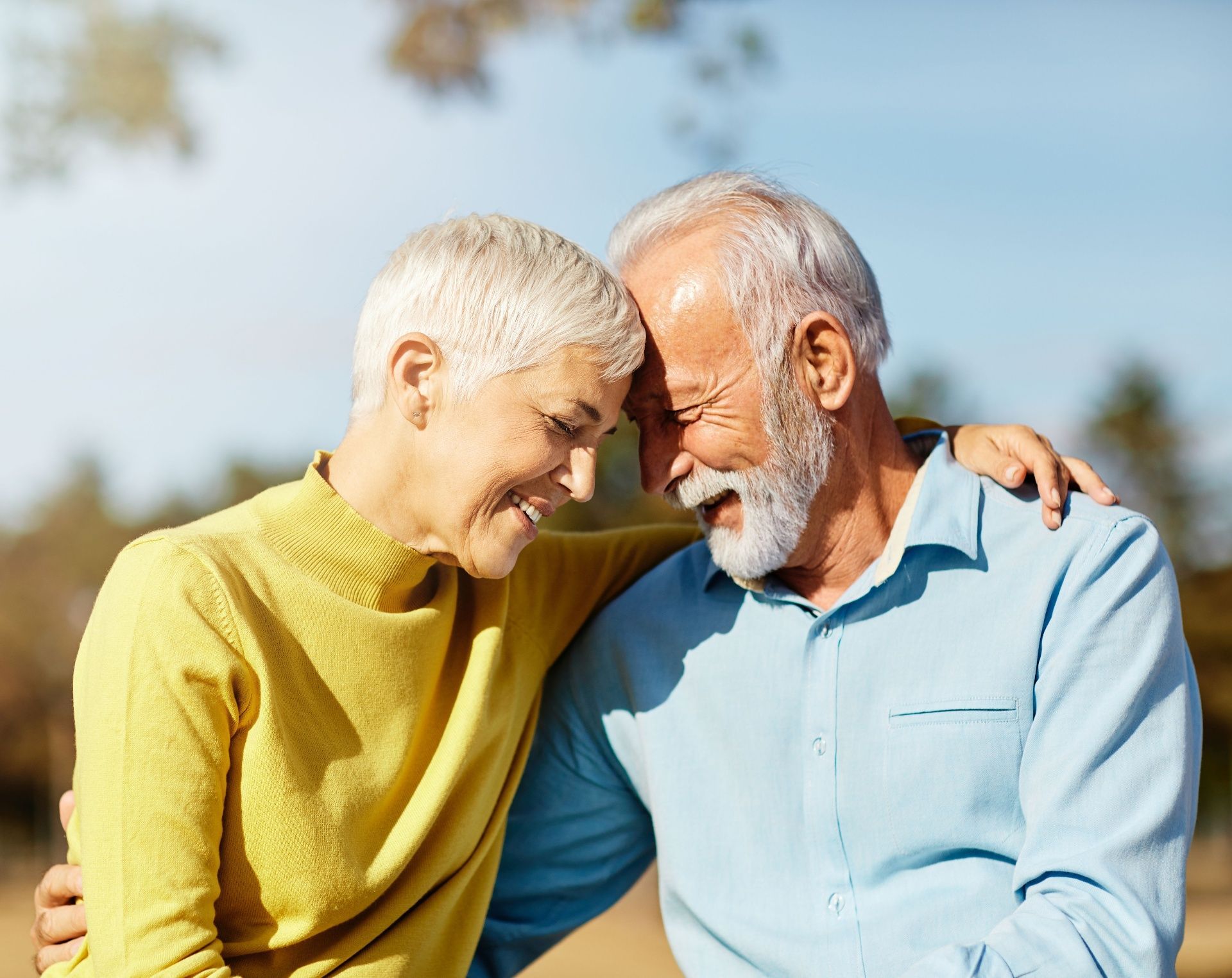 Two older adults smiling and leaning together outdoors on a sunny day