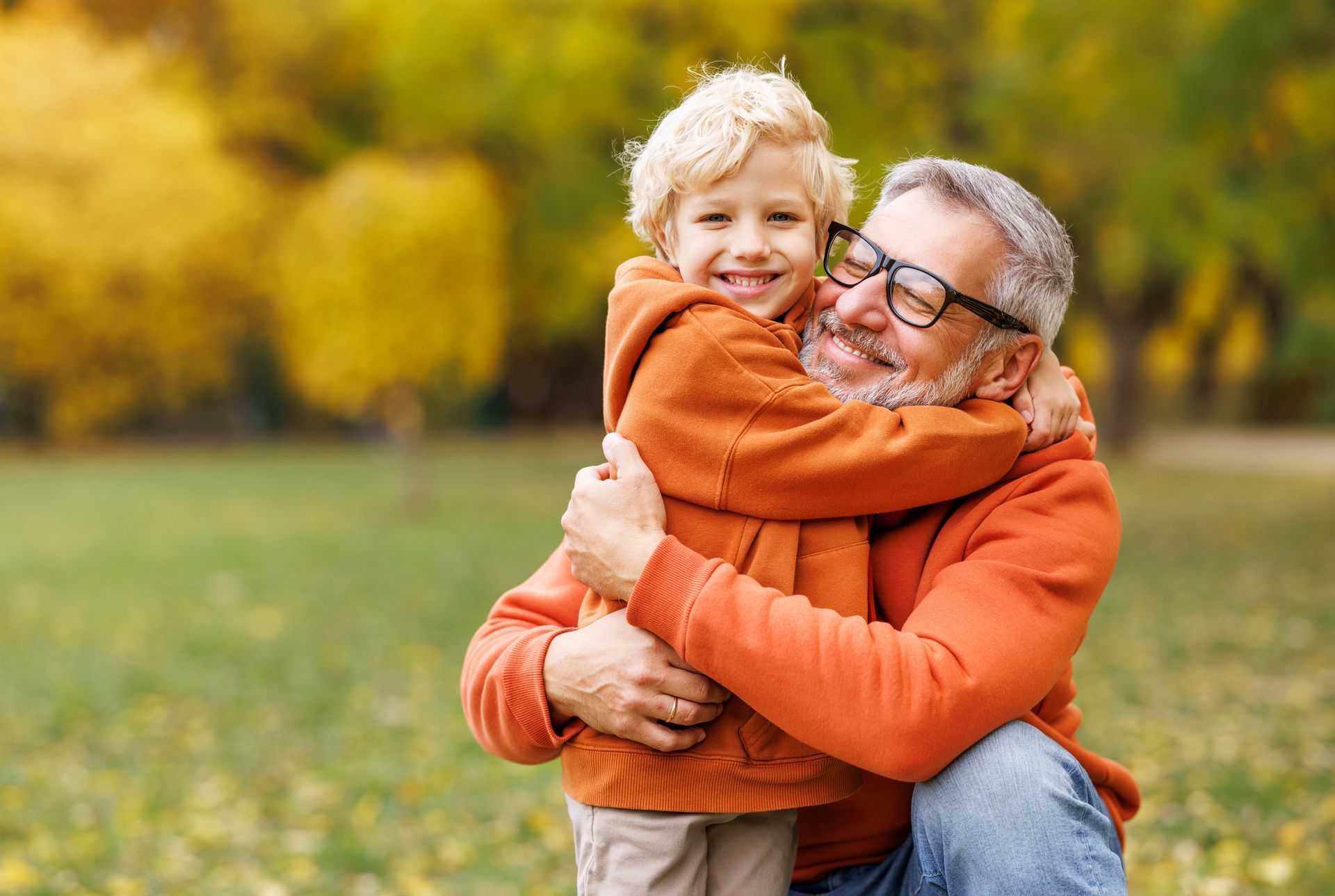 Smiling older man hugging a young child outdoors in autumn, with orange sweaters and blurred yellow trees.