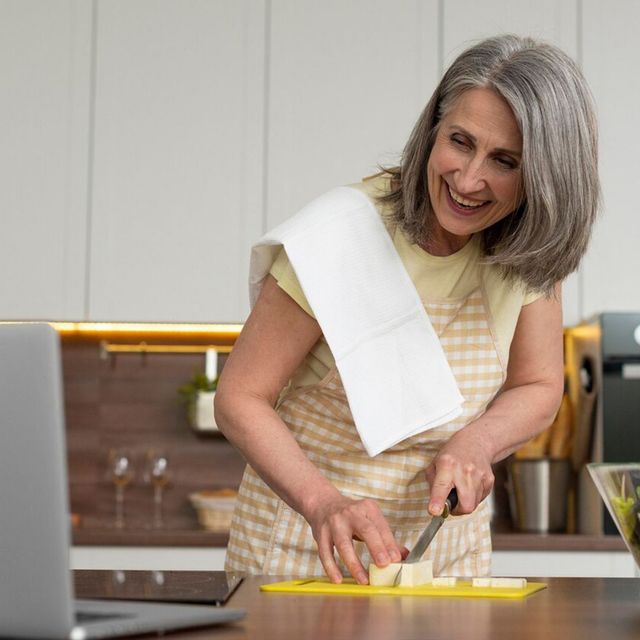 A woman is cutting a piece of cheese on a cutting board in front of a laptop.