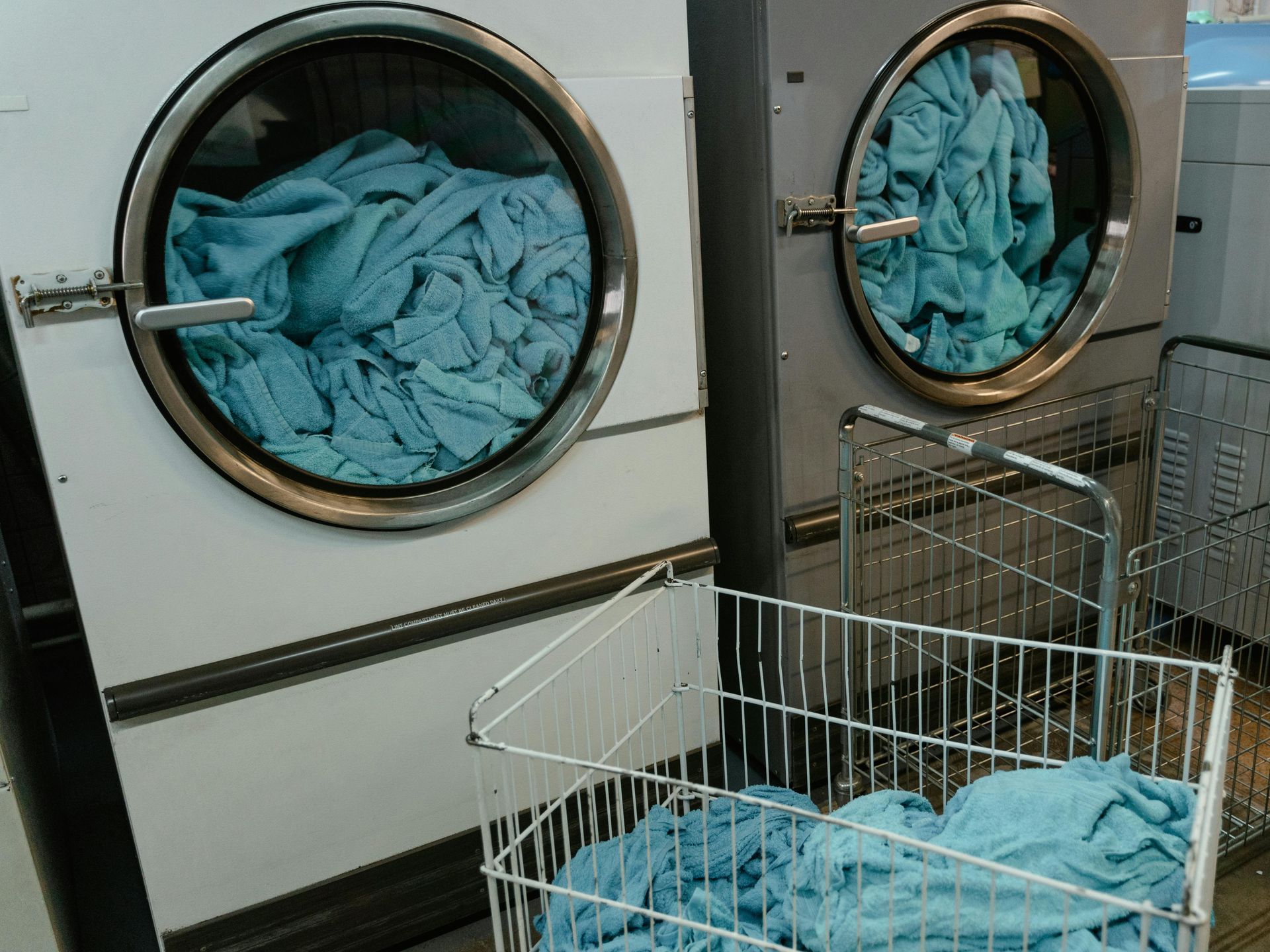Two industrial dryers containing blue towels stand behind a laundry cart filled with similar blue towels.
